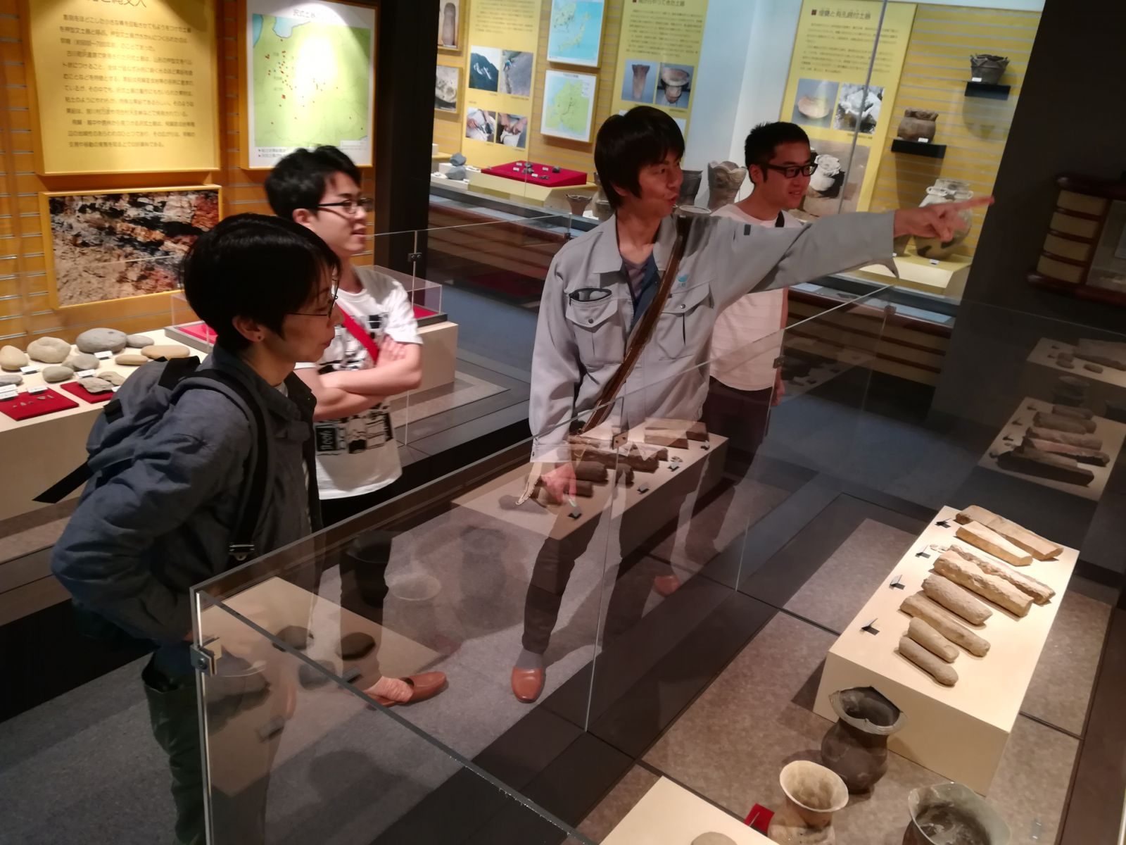 展示ケースの石棒を指さして解説するメンバーと来館者